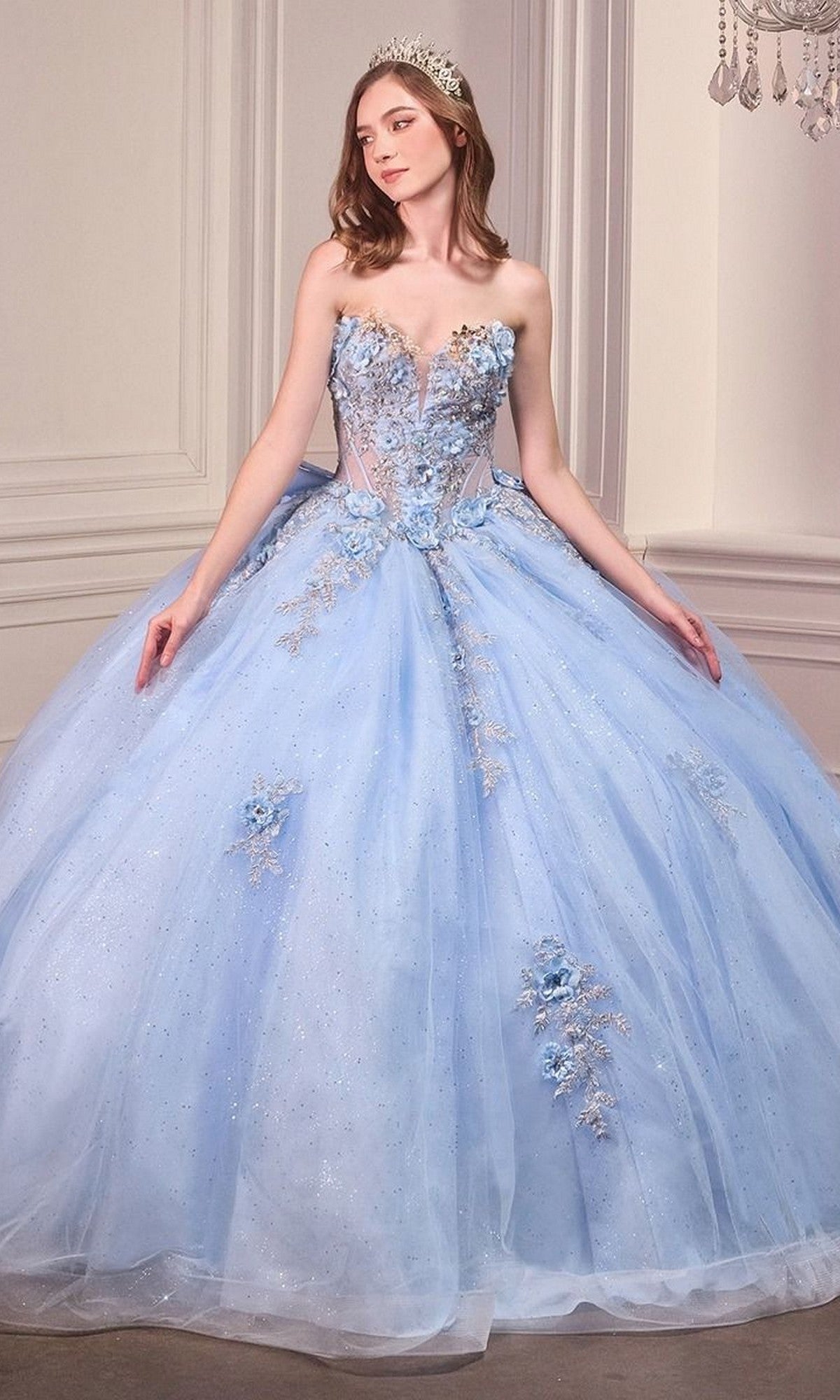 A woman wearing the Quinceanera Dress 15726 By Ladivine in light blue, featuring a strapless ball gown design with 3D flowers and a tiara, stands in an elegant room with pale walls.